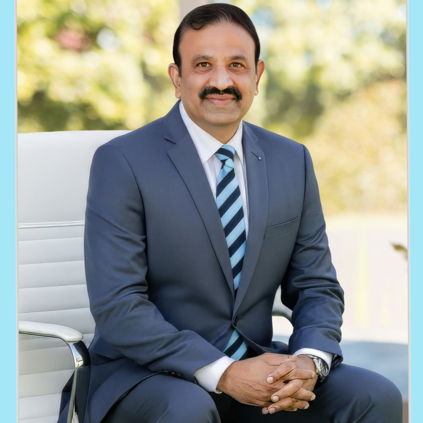 Smiling man with a mustache wearing a blue suit and striped tie, sitting in a chair.